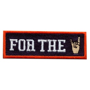 May include: An embroidered patch with an orange border and a dark blue background. The patch says "FOR THE" in white lettering with a gold-coloured rock and roll hand gesture to the right of the text.