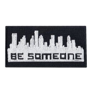 May include: White embroidered patch on a black background with a cityscape silhouette and the text "BE SOMEONE".
