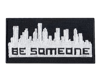 May include: White embroidered patch on a black background with a cityscape silhouette and the text "BE SOMEONE".