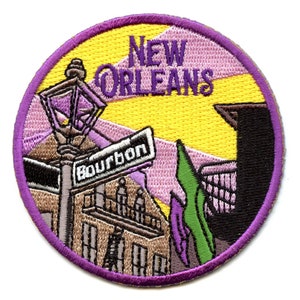 Op de afbeelding: Een geborduurde patch in paars en geel met een straatbeeld van New Orleans. De patch toont een lantaarnpaal, een gebouw met een balkon en een bord met de tekst "Bourbon".
