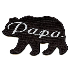 May include: A black embroidered patch in the shape of a bear with the word "Papa" in white cursive script. The patch is designed to be ironed or sewn onto fabric, suitable for clothing or accessories.