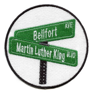 May include: A round, white patch with a black border. The patch features a green street sign with the text "Belfort Ave" and another green street sign with the text "Martin Luther King Blvd".