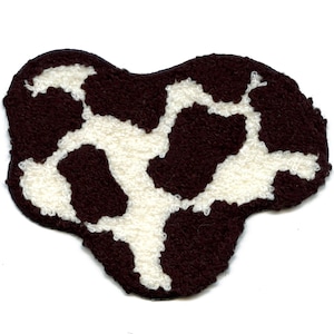 May include: A brown and white cow print rug with a unique shape. The rug features a dark brown background with irregular white patches, creating a distinctive pattern. The rug is made of a soft, textured material.