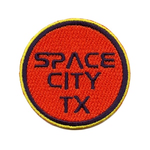 Könnte beinhalten: Ein runder, orangefarbener gestickter Aufnäher mit schwarzem Rand und gelber Umrandung. Der Text "SPACE CITY TX" ist in Lila gestickt.