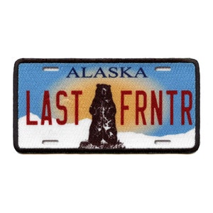 May include: A blue and white Alaska license plate patch with a brown bear standing on a snowy background. The text on the plate reads "LAST FRNTR".