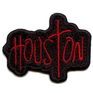Può includere: Una toppa ricamata nera con la scritta "Houston" in rosso. La toppa ha una forma unica e un design a croce rossa incorporato nella scritta.