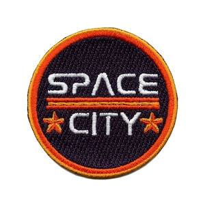 May include: A round, black embroidered patch with an orange border. The patch says "SPACE CITY" in white text with two orange stars on either side of the text.