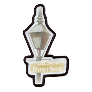 May include: Embroidered patch featuring a stylized street lamp and a sign that reads "Rue Bourbon" and "Bourbon" in a serif font. The patch has a white background with a brown border.
