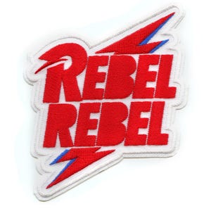 Könnte beinhalten: Roter und blauer gestickter Aufnäher mit dem Text "Rebel Rebel" in fetter Schrift. Der Aufnäher zeigt ein Blitz-Design.