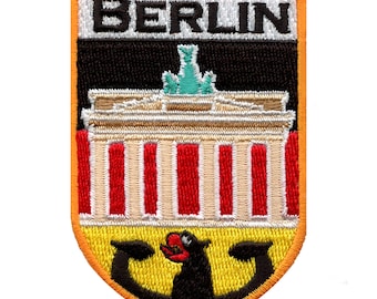 May include: An embroidered patch featuring the word "BERLIN" in white lettering on a black background. The patch depicts a stylised image of the Brandenburg Gate in red, white and black, with a yellow and black bear at the bottom.