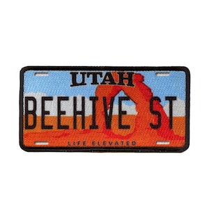Könnte beinhalten: Ein schwarz-weißes Patch mit einem roten und orangefarbenen Bild des Beehive State, dem Spitznamen für Utah. Das Patch trägt den Text "UTAH BEEHIVE ST" und "LIFE ELEVATED" in Schwarz.