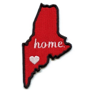 Pode incluir: Etiqueta bordada vermelha em forma do estado do Maine com a palavra "home" e um coração branco no canto inferior esquerdo.