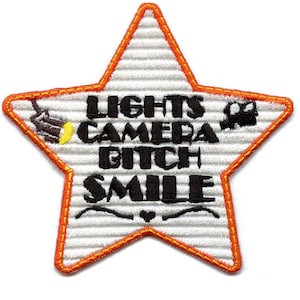 May include: Embroidered star-shaped patch with the text "LIGHTS CAMERA BITCH SMILE". The patch has a white background with horizontal lines, an orange border, and black text. Includes illustrations of a film camera and a spotlight.