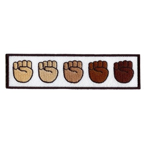 May include: Five raised fists in a row, each a different shade of brown, from light to dark. The fists are embroidered on a white background with a brown border.