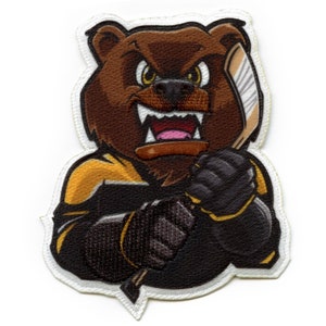 May include: A brown bear mascot wearing a black and yellow hockey jersey and holding a hockey stick. The bear has a fierce expression with sharp teeth and angry eyes.