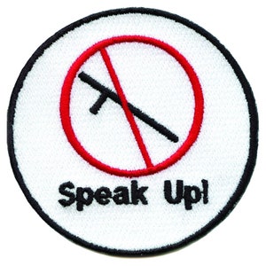 Op de afbeelding: Geborduurde patch met een witte achtergrond, een zwarte rand en de woorden "Speak Up!" in het zwart. Het ontwerp bevat een rode cirkel met een zwart object doorgestreept, wat een verbod symboliseert.