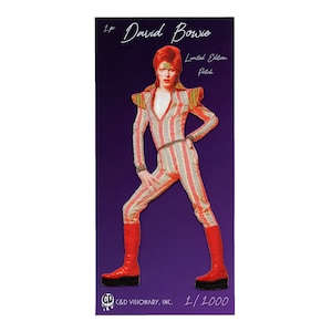 May include: A red, white and yellow striped jumpsuit with a red and white striped collar. The figure is wearing red platform shoes and has red hair. The image is a limited edition patch with the text "David Bowie" and "Limited Edition Patch" printed on it. The patch is numbered 1/1000.