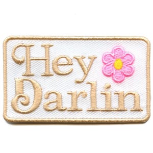 May include: Embroidered patch with the words "Hey Darlin" in a tan script font. A pink flower with a yellow center is on the right. The patch has a white background and a tan border.