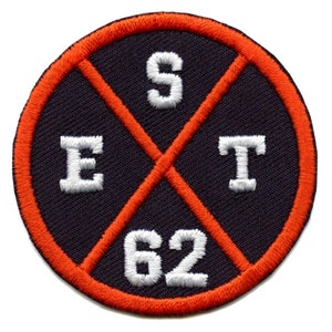 May include: A round, black patch with an orange border. The patch has a white embroidered cross with the letters "EST" and "62" in the center.