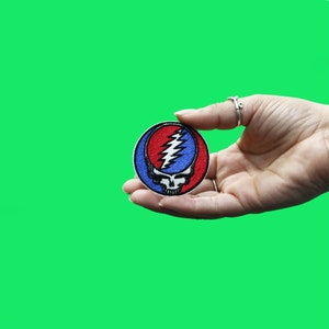 May include: A red, white, and blue embroidered patch featuring a skull with a lightning bolt design. The patch is held in a hand.
