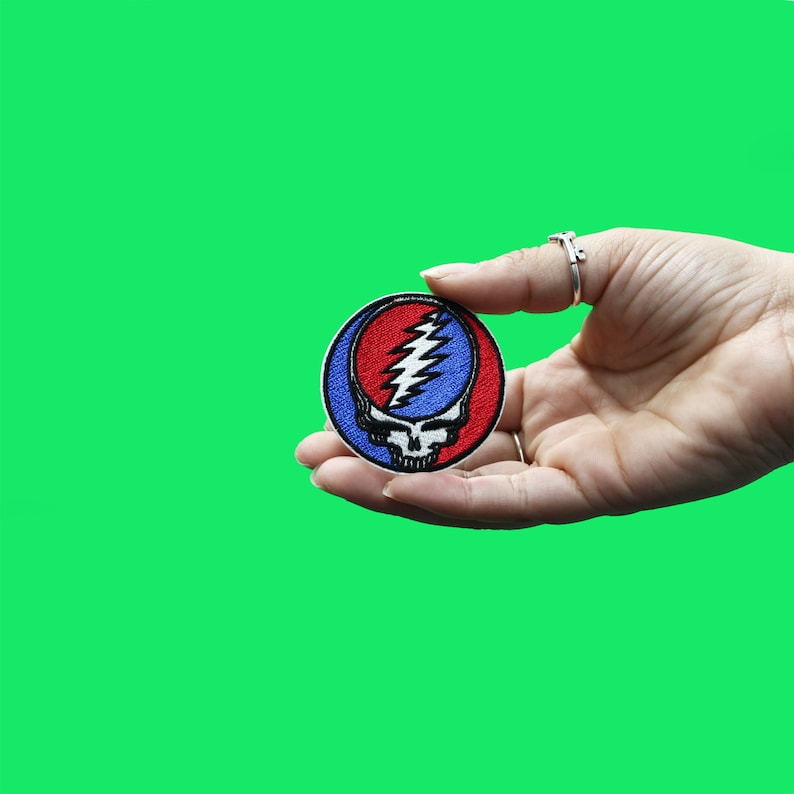 May include: A red, white, and blue embroidered patch featuring a skull with a lightning bolt design. The patch is held in a hand.