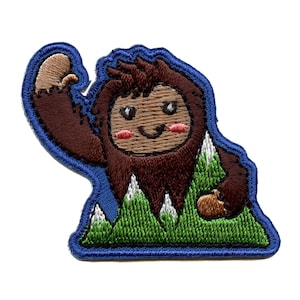 Op de afbeelding: Geborduurde patch met een bruine cartoon Bigfoot met een vriendelijk gezicht, staand in een groen berglandschap met witte sneeuwtoppen. De patch heeft een blauwe rand.