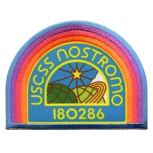 Op de afbeelding: Een blauwe en gele geborduurde patch met een regenboogrand. De patch heeft een gestileerde afbeelding van een planeet met een ster en de tekst "USCSS Nostromo 180286".