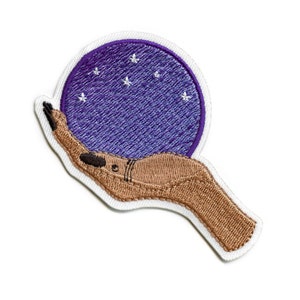 May include: An embroidered patch of a hand holding a purple orb with white stars. The hand is brown and the orb is surrounded by a white outline.