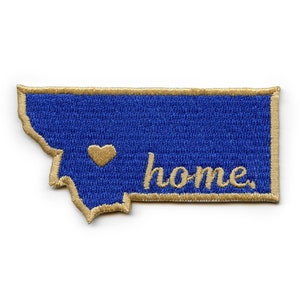 May include: A blue embroidered patch in the shape of the state of Montana with a gold outline. The patch has a gold heart in the center and the word "home" embroidered in gold below the heart.