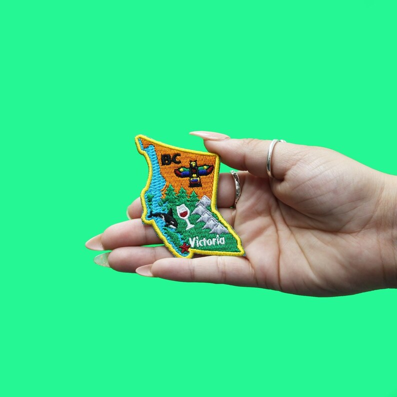 May include: A yellow, green, and blue embroidered patch shaped like the province of British Columbia, Canada. The patch features the text "BC" and "Victoria" and includes an image of a totem pole, a whale, a glass of wine, and mountains.
