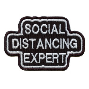 Könnte beinhalten: Schwarzer und weißer gestickter Aufnäher mit dem Text "SOCIAL DISTANCING EXPERT" in einem weißen Umriss.