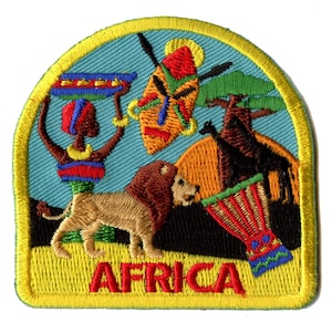Op de afbeelding: Een geborduurde patch in geel, blauw en oranje met een leeuw, een vrouw die een kom vasthoudt, een masker en een trommel. De tekst "AFRICA" is in rood onderaan de patch geborduurd.