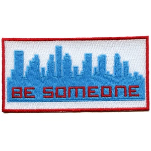 May include: A white embroidered patch with a red border. The patch features a blue cityscape silhouette and the text "BE SOMEONE" in red.
