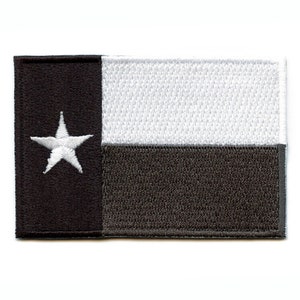 May include: A black and white embroidered patch featuring the Texas state flag. The flag has a single white star on a black background, with a white stripe and a dark gray stripe below.