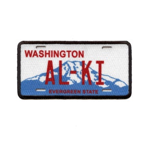 May include: Embroidered patch resembling a Washington state license plate. The patch features the word "WASHINGTON" above the red letters "ALKI" and a blue mountain range. The text "EVERGREEN STATE" is below the mountain.