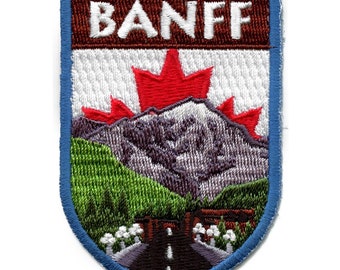 May include: An embroidered patch with a blue border, a white background, and a red maple leaf. The patch features a mountain scene with a road and the word "BANFF" in brown letters.