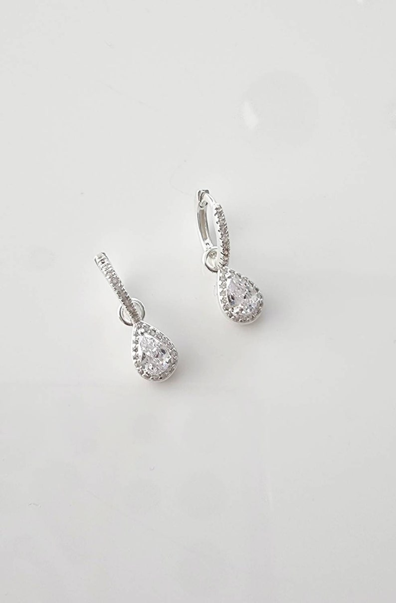 Pu&ograve; includere: Un paio di orecchini a cerchio d'argento con un pendente a forma di pera in pietra trasparente. La pietra &egrave; circondata da pietre trasparenti pi&ugrave; piccole.