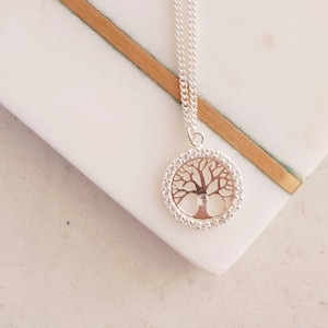 Puede incluir: Un collar de plata con un colgante de árbol de la vida. El colgante es redondo y tiene un cristal transparente en el centro. La cadena es delicada y tiene un acabado plateado.