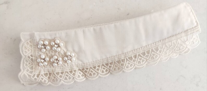 Puede incluir: Un cuello de tela blanca con ribete de encaje y adornos de perlas.