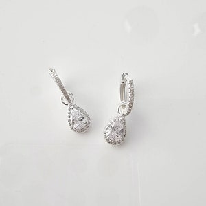 Pu&ograve; includere: Un paio di orecchini a cerchio d'argento con una pietra trasparente a forma di pera che pende da ogni cerchio.