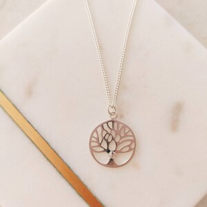 Puede incluir: Un collar de plata con un colgante de árbol de la vida. El colgante es un círculo con un diseño de árbol detallado y una pequeña piedra redonda en el centro.