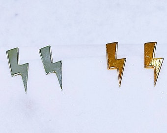 Puede incluir: Dos pares de pendientes de botón con forma de rayo, uno plateado y otro dorado. Los pendientes tienen un diseño minimalista. Se muestran sobre una superficie blanca. Cada pendiente mide aproximadamente 1 cm de largo.