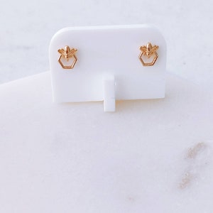 Puede incluir: Un par de pendientes de botón dorados con un diseño de abeja pequeña sobre un contorno hexagonal de panal. Los pendientes están montados en una tarjeta de presentación blanca y presentan un acabado metálico pulido y alas detalladas.