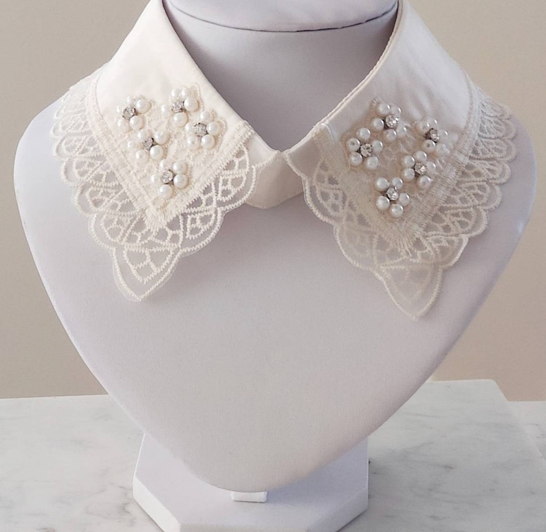 Puede incluir: Un collar de encaje blanco con adornos de perlas y pedrer&iacute;a. El collar est&aacute; en un busto de maniqu&iacute; blanco.