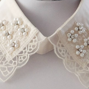 Puede incluir: Un cuello de encaje blanco con adornos de perlas y pedrer&iacute;a. El cuello tiene un borde festoneado y est&aacute; dise&ntilde;ado para ser usado sobre una camisa o blusa.