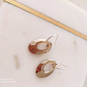 Puede incluir: Un par de pendientes colgantes de color dorado con un diseño ovalado facetado. Los pendientes tienen un cierre de gancho simple.