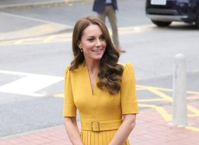 Pu&ograve; includere: Una donna con un vestito giallo con scollo a V e cintura. Ha i capelli lunghi e castani e sorride.