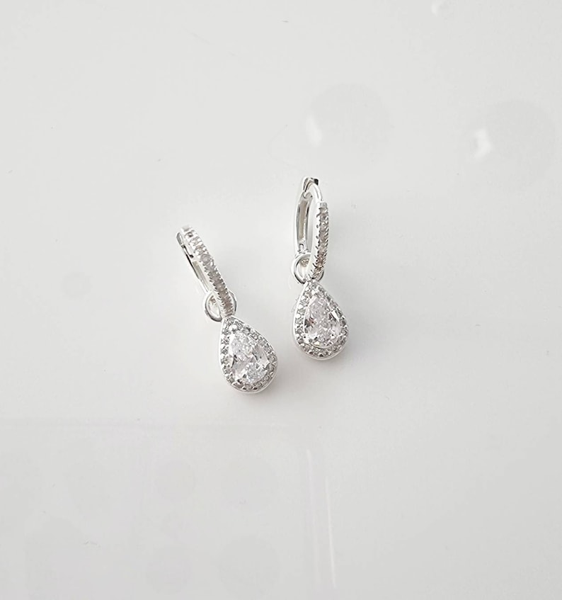 Pu&ograve; includere: Un paio di orecchini a cerchio d'argento con un ciondolo di cristallo a forma di pera. Il cristallo &egrave; circondato da cristalli pi&ugrave; piccoli.