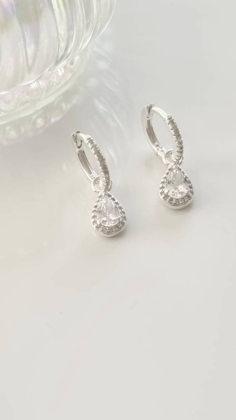 Pu&ograve; includere: Un paio di orecchini a cerchio d'argento con un ciondolo di cristallo a forma di lacrima. Il cristallo &egrave; circondato da un'aureola di cristalli pi&ugrave; piccoli.