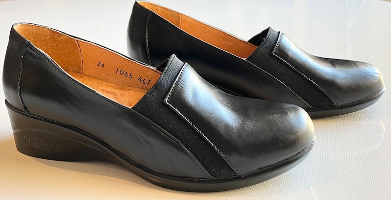 Peut inclure: Une paire de chaussures compens&eacute;es en cuir noir avec un panneau &eacute;lastique noir sur le dessus. Les chaussures ont une semelle int&eacute;rieure en cuir marron avec les num&eacute;ros 24, 1065 et 467 imprim&eacute;s dessus.
